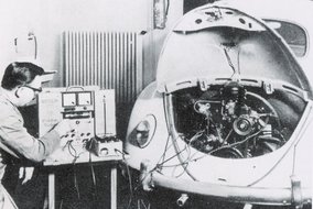  Schwarz-weiß Foto einer historischen Fahrzeugprüfung mit Messgeräten und einem VW Käfer in einer Werkstatt.