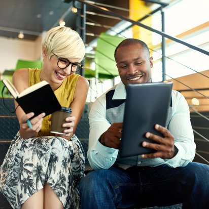 Zwei Personen, eine mit einem Notizbuch und die andere mit einem Tablet, sitzen zusammen auf einer Treppe und arbeiten