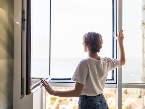 Eine Person öffnet ein Fenster und schaut hinaus. Sie trägt ein weißes T-Shirt und eine Jeans, während sie von hinten zu sehen ist.