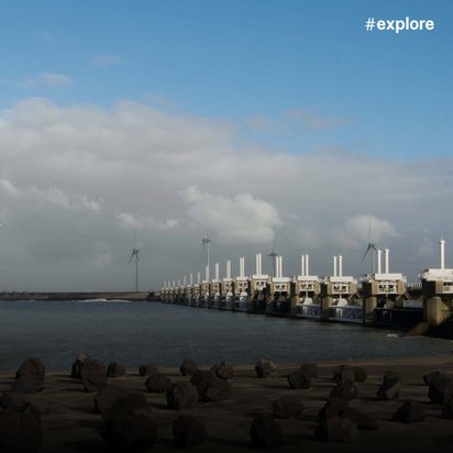 Eine massive Sturmflutschutzanlage mit weißen Sperrwerken erstreckt sich über das Wasser, dahinter drehen sich Windkraftanlagen. Die Küstenschutzbarriere mit ihren charakteristischen Toren schützt das Hinterland vor Sturmfluten bei blau-bewölktem Himmel.