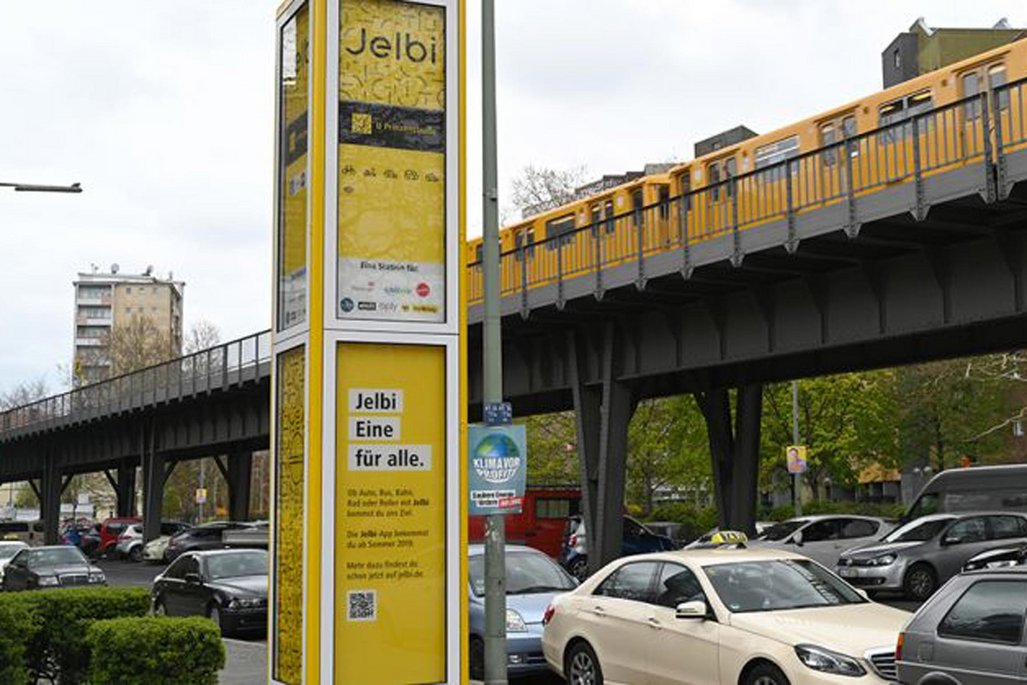Gelbe Werbetafel mit "Jelbi" Branding vor einer Straßenszene mit parkenden Autos und einer Bahnbrücke im Hintergrund.