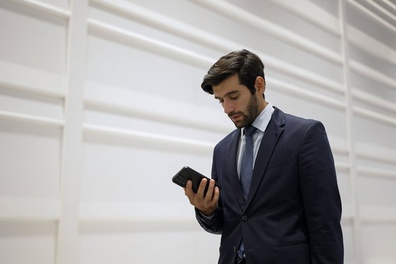 Geschäftsmann im Anzug blickt konzentriert auf sein Smartphone vor heller Wand