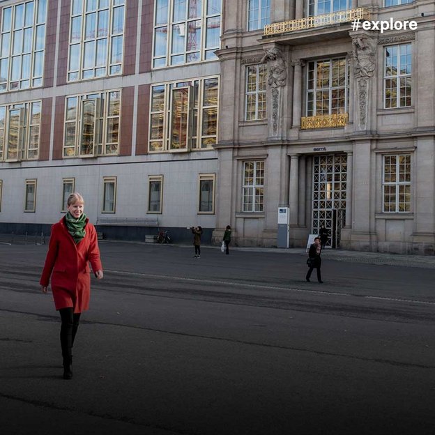 Eine Person in rotem Mantel und grünem Schal geht vor einem großen, repräsentativen Gebäude mit klassischer Fassade und modernen Anbauten über einen Platz. Weitere Menschen sind im Hintergrund zu sehen.