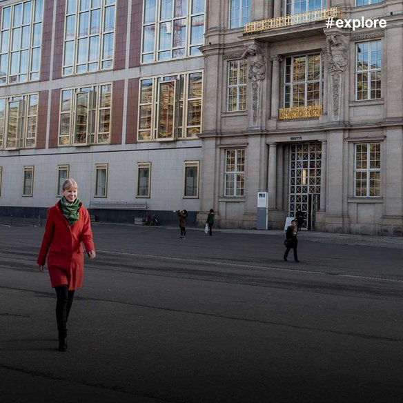 Eine Person in rotem Mantel und grünem Schal geht vor einem großen, repräsentativen Gebäude mit klassischer Fassade und modernen Anbauten über einen Platz. Weitere Menschen sind im Hintergrund zu sehen.