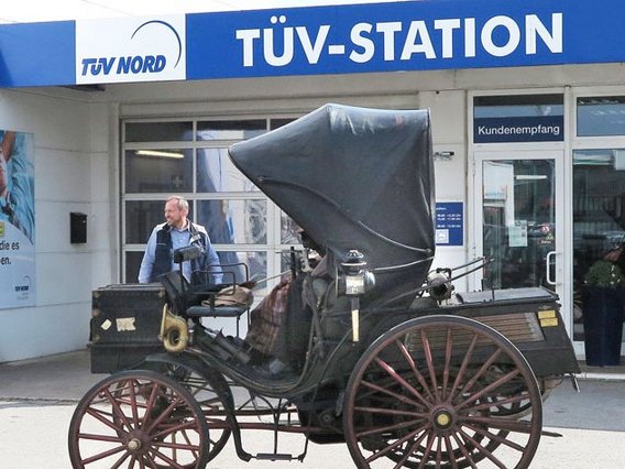 Historische Pferdekutsche vor einer modernen TÜV-Station, die den Wandel der Verkehrstechnik symbolisiert.