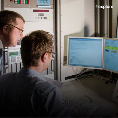zwei Wissenschaftler oder Techniker in einem Kontrollraum, die auf zwei Computerbildschirme mit Daten und Diagrammen schauen. Durch eine Glasscheibe beobachten sie komplexe Laborausrüstung mit Röhren und technischen Instrumenten.