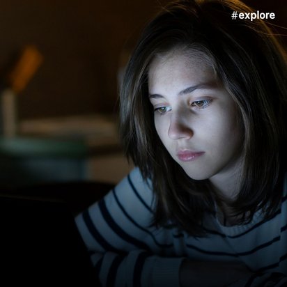 Junge Frau mit braunen Haaren im gestreiften Pullover arbeitet konzentriert am Laptop in dunklem Raum, beleuchtet vom Bildschirmschein.