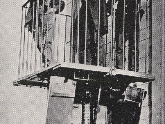 Historische Schwarz-Weiß-Fotografie von drei Personen, die auf einer Aufzugsplattform oder einem Baulift stehen, der an der Außenseite eines Gebäudes befestigt ist.