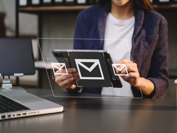Person hält Tablet mit schwebenden E-Mail-Symbolen, Laptop und Kaffeebecher im Hintergrund.