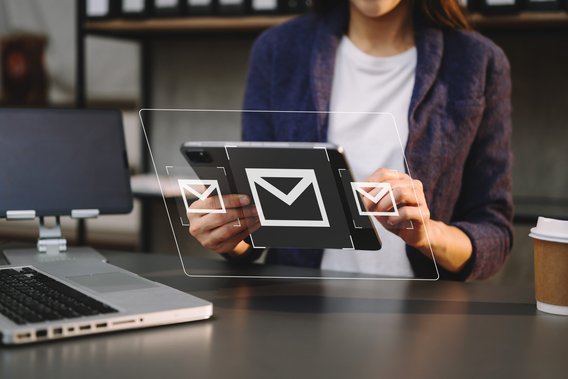 Person hält Tablet mit schwebenden E-Mail-Symbolen, Laptop und Kaffeebecher im Hintergrund.