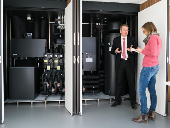 Ein Technikraum mit geöffneten Schranktüren zeigt schwarze Heizungs- oder Klimageräte. Rechts steht ein Mann im Anzug im Gespräch mit einer Frau, die Notizen macht.