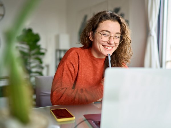  Junge Frau mit Brille und rotem Pullover sitzt lächelnd vor einem Laptop am Schreibtisch