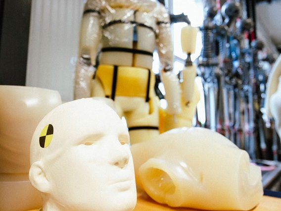 Nahaufnahme von Dummy-Köpfen mit Crashtest-Markierungen im Vordergrund, dahinter hängende Test-Dummies.