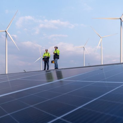 Zwei TÜV NORD-Ingenieure prüfen Solarmodule vor Windrädern unter blauem Himmel