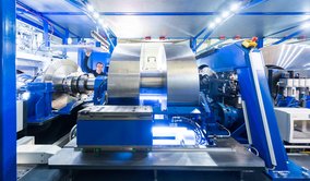  In einer hochmodernen industriellen Anlage ist eine große, komplexe Maschine zu sehen, die aus blauen und silbernen Komponenten besteht. Im Zentrum der Maschine befindet sich ein massives, zylindrisches Metallteil, das von verschiedenen mechanischen Armen und Halterungen umgeben ist. Die Umgebung ist hell erleuchtet, mit zahlreichen Lichtern, die die Arbeitsfläche ausleuchten.