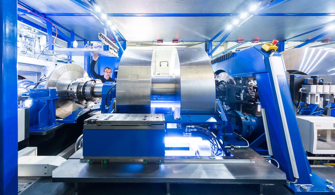  In einer hochmodernen industriellen Anlage ist eine große, komplexe Maschine zu sehen, die aus blauen und silbernen Komponenten besteht. Im Zentrum der Maschine befindet sich ein massives, zylindrisches Metallteil, das von verschiedenen mechanischen Armen und Halterungen umgeben ist. Die Umgebung ist hell erleuchtet, mit zahlreichen Lichtern, die die Arbeitsfläche ausleuchten.