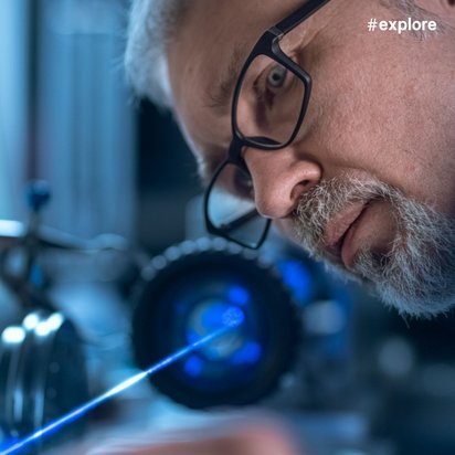 Nahaufnahme eines älteren Mannes mit Brille und grauem Bart, der konzentriert auf einen blauen Laserstrahl in einem wissenschaftlichen Labor blickt, umgeben von optischen Geräten.