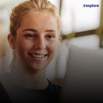 Eine junge Frau mit blonden Haaren lächelt in einem Innenraum mit unscharfem Hintergrund, vermutlich in einem Büro oder Bildungsumfeld.