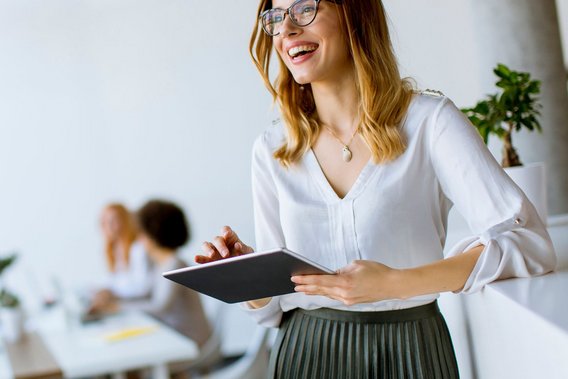 Eine Frau mit Brille lächelt und hält ein Tablet in den Händen, im Hintergrund arbeiten weitere Personen im Büro.