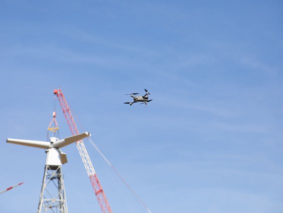 Ein Drohne fliegt vor blauem Himmel neben einem weißen Windkraftanlagen-Turm mit rotem Kran im Hintergrund. Die Drohne führt vermutlich eine Inspektion der Windkraftanlage durch.