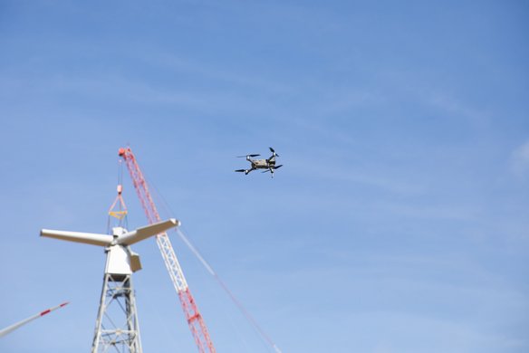 Ein Drohne fliegt vor blauem Himmel neben einem weißen Windkraftanlagen-Turm mit rotem Kran im Hintergrund. Die Drohne führt vermutlich eine Inspektion der Windkraftanlage durch.