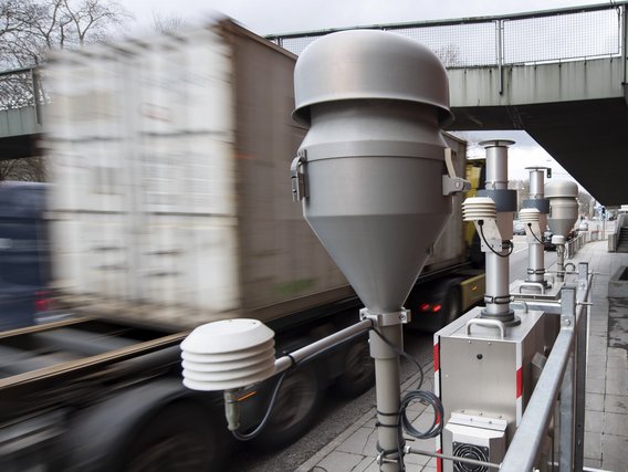 Eine Reihe von grauen Luftqualitätsmessgeräten steht entlang einer Bahnstrecke, während ein unscharfer Zug vorbeifährt. Die Messstation erfasst Umweltdaten und ist mit verschiedenen Sensoren und Geräten ausgestattet.