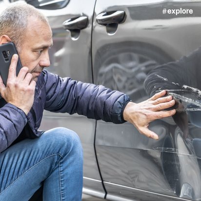 Ein Mann kniet neben einem grauen Auto mit Kratzern, spricht am Telefon und berührt die Kratzer am Auto.
