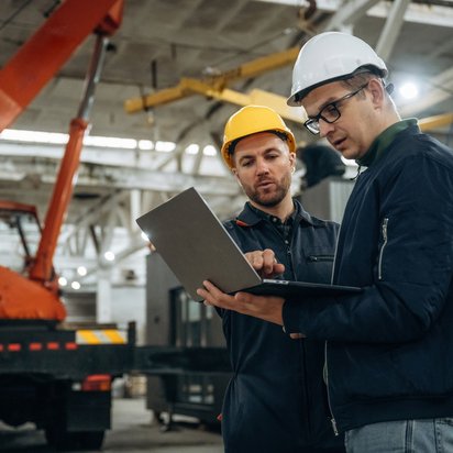 Zwei Ingenieure mit Schutzhelmen prüfen Produktionsdaten auf einem Laptop in der Werkshalle
