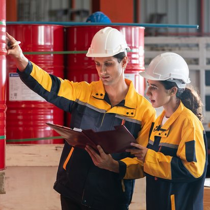 Sicherheitsprüfung im Gefahrstofflager Zwei Fachkräfte mit Schutzkleidung und Helm prüfen rote Fässer in einem Lager