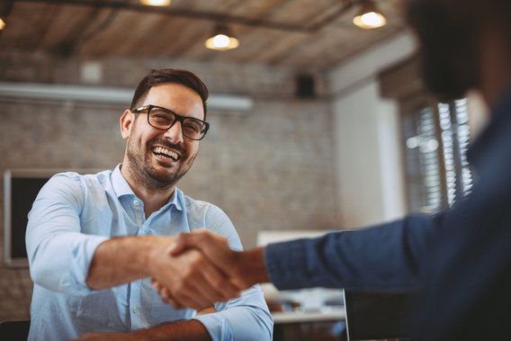 Ein Mann mit Brille und Hemd lacht und schüttelt einer anderen Person im Büro die Hand.