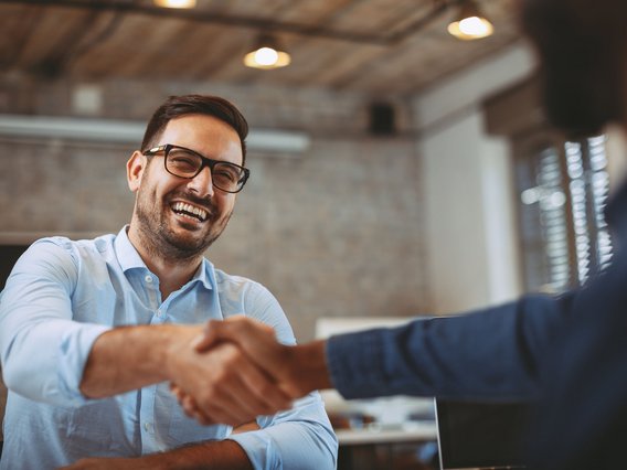 Ein Mann mit Brille und Hemd lacht und schüttelt einer anderen Person im Büro die Hand.