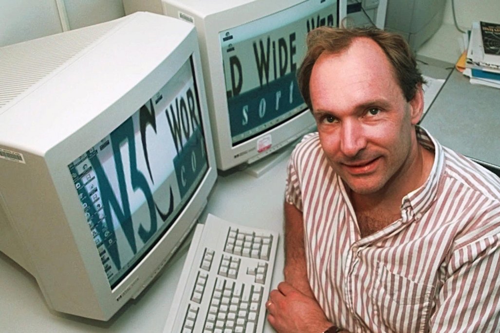 Tim Berners-Lee in gestreiftem Hemd sitzt vor einem Computer mit CRT-Monitor, auf dem "World Wide Web" zu lesen ist, typische 1990er Jahre Büroeinrichtung.