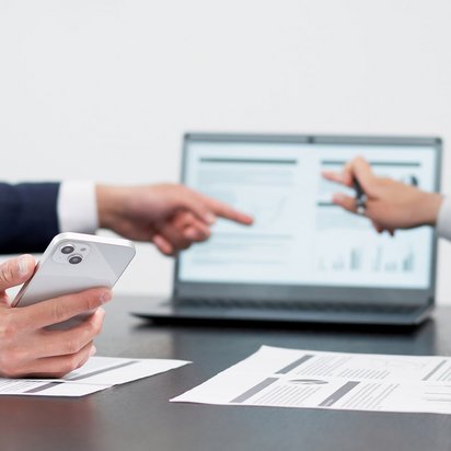 Teammeeting zur Auswertung von Geschäftszahlen mit digitalen und analogen Tools Zwei Personen zeigen auf Diagramme auf einem Laptop, im Vordergrund ein Smartphone und ausgedruckte Unterlagen