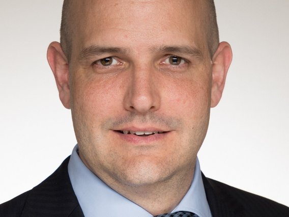 Martin Saalmann trägt einen schwarzen Anzug mit hellblauem Hemd und karierter Krawatte. Er lächelt leicht und blickt direkt in die Kamera vor einem neutralen Hintergrund.
