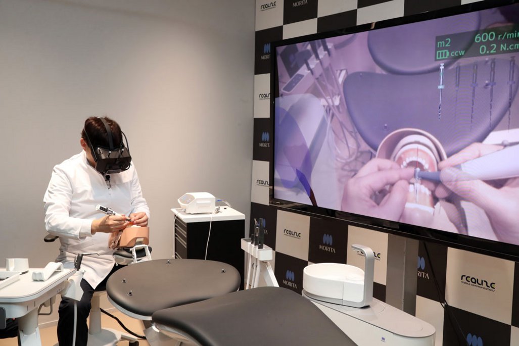 Ein medizinischer Simulationsraum mit einer Person in weißem Kittel und VR-Brille, die an einem Übungsmodell arbeitet. Auf einem großen Bildschirm wird ein zahnmedizinischer Eingriff gezeigt.