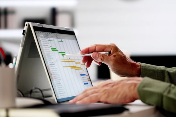 Hand zeigt mit Stift auf ein Gantt-Diagramm zur Projektplanung auf einem Laptop-Bildschirm.