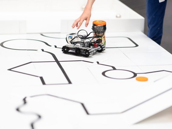 Ein kleiner Roboter mit Raupenantrieb fährt auf einer weißen Oberfläche, die mit schwarzen Linien markiert ist. Eine Hand greift nach dem Roboter, der einen orangefarbenen Behälter trägt. Es handelt sich um eine Übungssituation für Linienfolge-Roboter.
