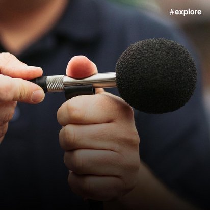 In der Nahaufnahme hält eine Person ein schwarzes Mikrofon in der rechten Hand und blaue Audiokabel in der linken Hand. Die Aufnahme zeigt wahrscheinlich einen Techniker oder Reporter bei der Arbeit.