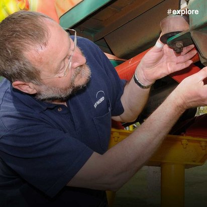 Ein Techniker in dunkelblauem Poloshirt inspiziert oder wartet die mechanischen Komponenten einer Fahrgeschäft-Anlage in einem Freizeitpark. Im Hintergrund sind bunte Elemente der Attraktion zu erkennen.