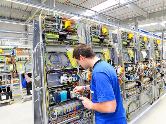 Elektrotechnische Arbeiten an Steuerungssystemen in der Fertigung. Techniker in blauer Kleidung arbeitet an einer industriellen Schalttafel in einer Fertigungshalle.