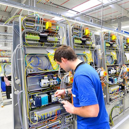  Techniker in blauer Kleidung arbeitet an einer industriellen Schalttafel in einer Fertigungshalle.