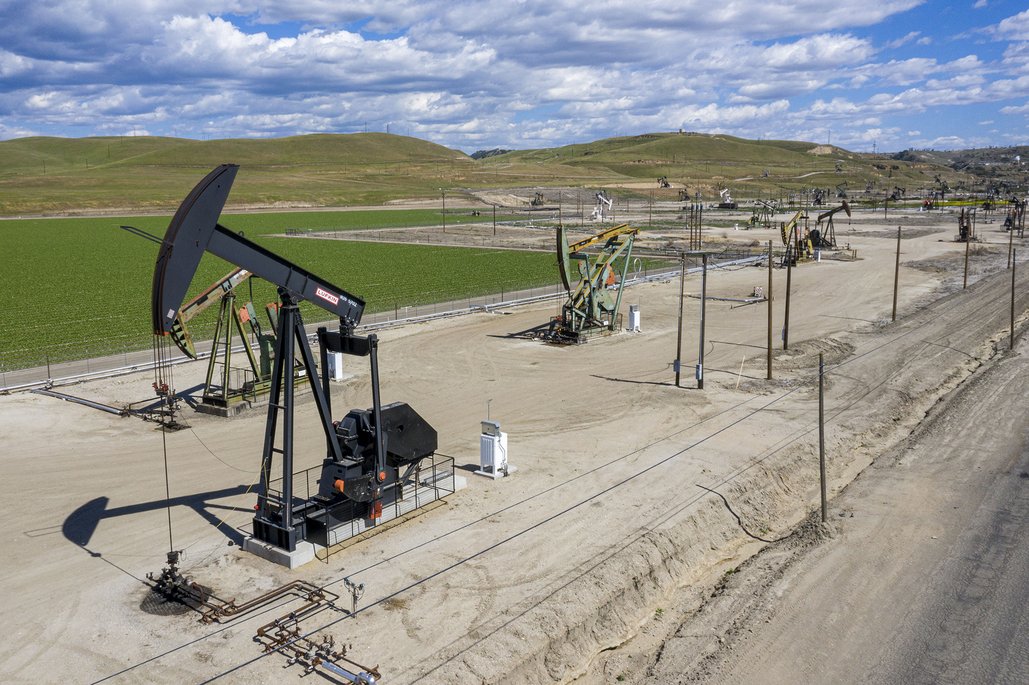 Luftaufnahme eines Ölfördergebiets mit mehreren schwarzen Pumpjacks (Pferdekopfpumpen) auf staubigen Betonflächen vor grünen Feldern und sanften Hügeln. Die industrielle Ölförderanlage erstreckt sich über eine weite Landschaft unter einem bewölkten blauen Himmel.