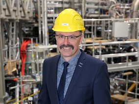 Robert Wolf mit Schnurrbart und gelbem Schutzhelm lächelt in einer technischen Industrieumgebung. Er trägt einen dunkelblauen Anzug und wirkt professionell.