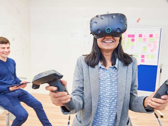 Eine Frau mit VR-Brille und Handheld-Controllern lächelt während einer Virtual-Reality-Erfahrung in einem modernen Büroraum, während ein Kollege im Hintergrund zuschaut.