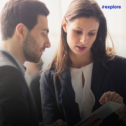 Zwei Geschäftsleute in Anzügen schauen gemeinsam auf ein Tablet oder Dokument, während im Hintergrund unscharf weitere Personen in einem hellen Büroraum zu sehen sind.