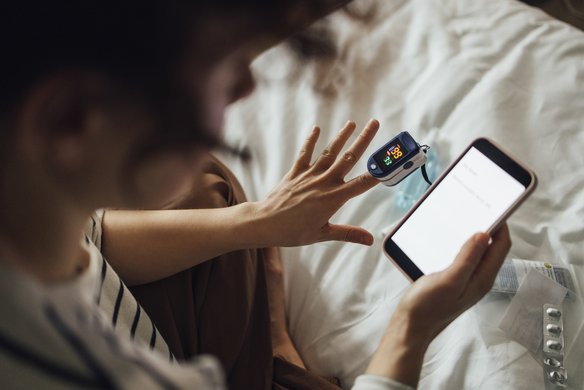 Person im Bett mit Pulsoximeter am Finger und Smartphone in der Hand, Medikamente liegen auf der Bettdecke.