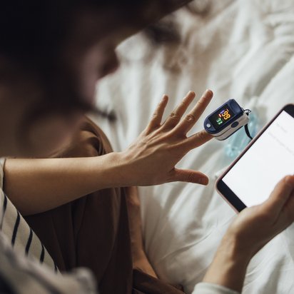 Person im Bett mit Pulsoximeter am Finger und Smartphone in der Hand, Medikamente liegen auf der Bettdecke.