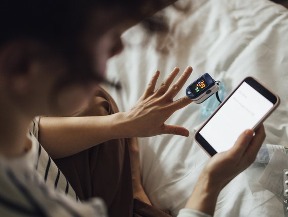 Person im Bett mit Pulsoximeter am Finger und Smartphone in der Hand, Medikamente liegen auf der Bettdecke.