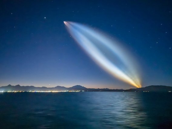 Ein heller Meteor mit langem Schweif erleuchtet den Nachthimmel über einem See mit Bergen im Hintergrund.