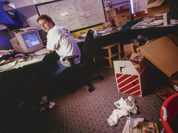 Ein Mann in weißem T-Shirt arbeitet an einem Computer in einem unordentlichen Büro mit Whiteboard und vielen Kartons.